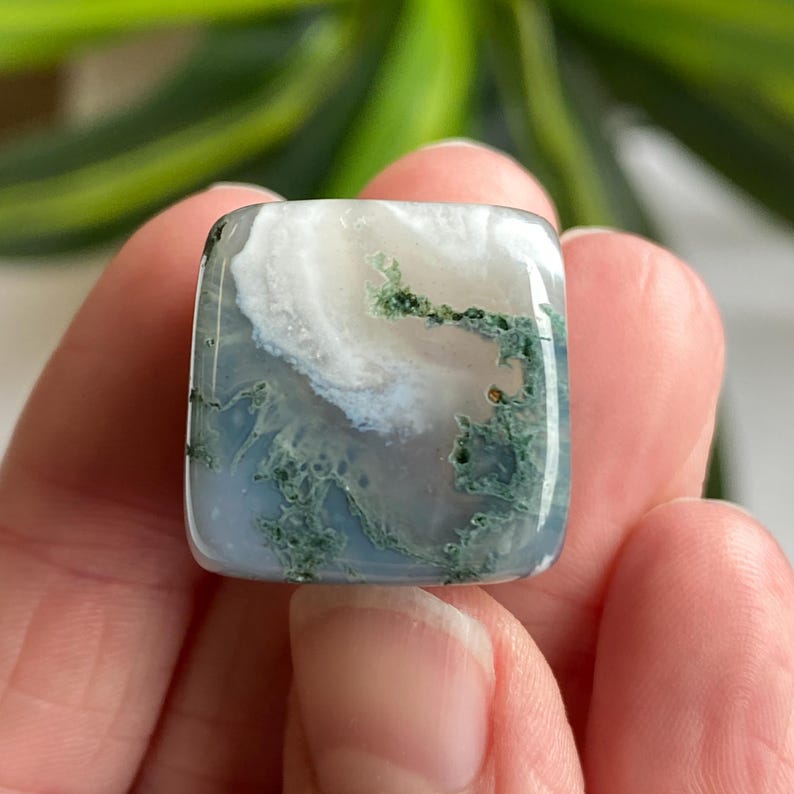 Puede incluir: Gema de &aacute;gata musgosa de forma cuadrada con superficie pulida. La piedra presenta una mezcla de colores blanco, azul y verde, con un patr&oacute;n arremolinado. La gema est&aacute; sujeta en una mano.