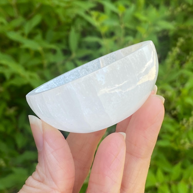 May include: A white selenite bowl, semi-circular in shape, held against a backdrop of green foliage. The bowl has a smooth, polished surface and a translucent quality, suggesting it is made of a crystal material. The bowl is likely used for decorative or spiritual purposes.