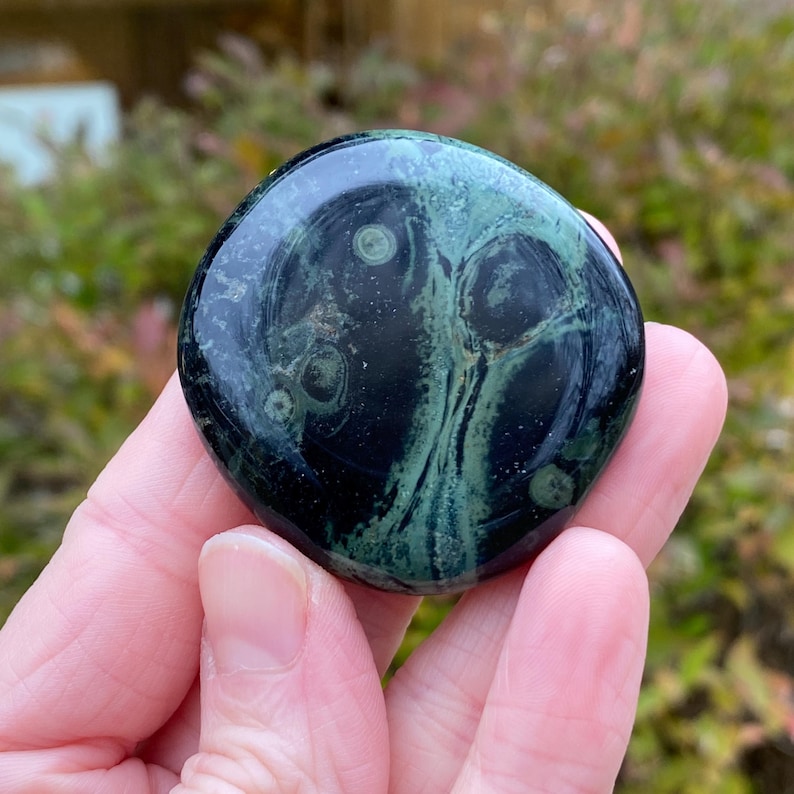 May include: A polished, round gemstone with a dark green and black swirling pattern. The stone has a glossy finish and features lighter green circular inclusions. The gemstone is held in a hand, with a blurred green background.
