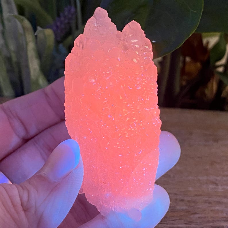 May include: A vibrant, glowing orange crystal cluster. The crystal has a rough, textured surface and a unique, irregular shape. The color is a bright, almost neon, orange, with hints of pink and yellow. The crystal is held in a hand.