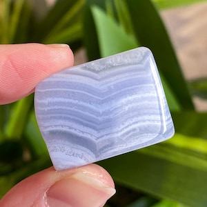 May include: A polished blue lace agate gemstone with a layered, banded pattern. The stone is a light blue color with white and darker blue bands. The gemstone is held between two fingers.