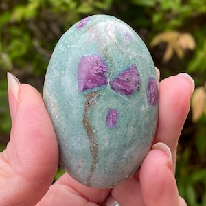 Może przedstawiać: Gładki, owalny kamień o turkusowej barwie podstawowej i żywych, magentowych wtrąceniach. Polerowany kamień ma naturalny wygląd. Magentowe wtrącenia są zgrupowane, tworząc unikalny wzór.