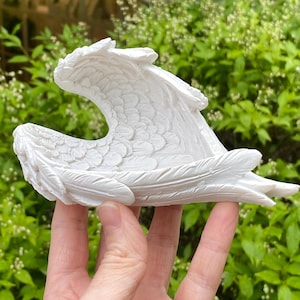May include: A white, sculpted decorative object shaped like a pair of feathered angel wings. The piece features detailed, layered plumage textures across the curved wing structure, creating a concave, bowl-like form with a smooth, matte finish.