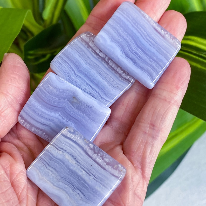 May include: Four square, polished blue lace agate stones with a banded pattern are held in a hand. The stones have a light blue color with darker blue and white striations. The background includes green and yellow leaves.