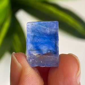 May include: A rectangular, translucent blue crystal with a textured surface. The crystal has shades of light and dark blue, with some white inclusions. The crystal is held between two fingers.