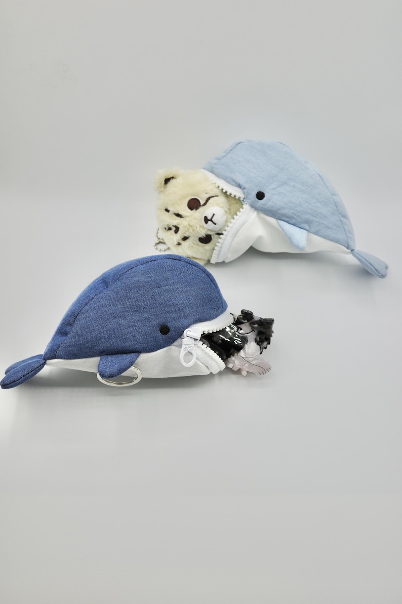 May include: Two plush whale-shaped zippered pouches. One is blue denim with a white lining and a silver key ring. The other is light blue with a white lining and a brown bear peeking out of the zippered opening.
