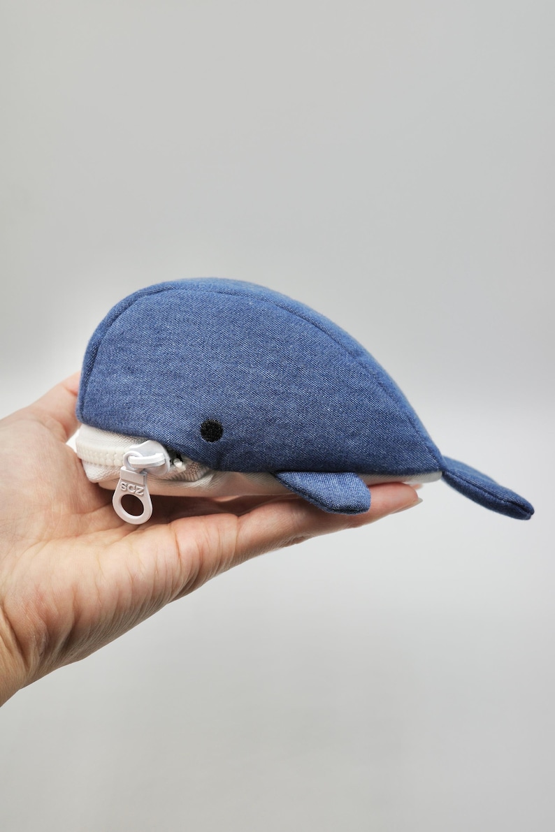 May include: A blue whale-shaped zippered pouch made of soft fabric. The pouch is held in a hand, and the zipper is visible.