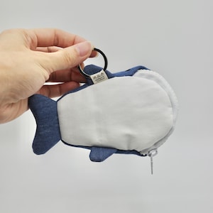 May include: A blue denim whale-shaped coin purse with a zipper closure and a metal ring attached. The purse has a white fabric lining and a small tag with text in a foreign language.