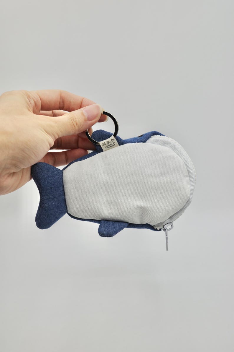 May include: A blue denim whale-shaped coin purse with a zipper closure and a metal ring attached. The purse has a white fabric lining and a small tag with text in a foreign language.