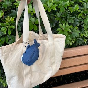 May include: A cream-coloured canvas tote bag with a blue whale plush keychain attached to the handle.