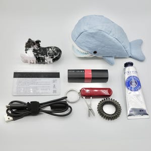 May include: A collection of various items including a black and white dog figurine, a blue whale plush toy, a black credit card, a black USB cable, a red lipstick, a red multi-tool keychain, a black hair tie, and a blue tube of hand cream labelled "L'OCCITANE CREME MAINS HAND CREAM".
