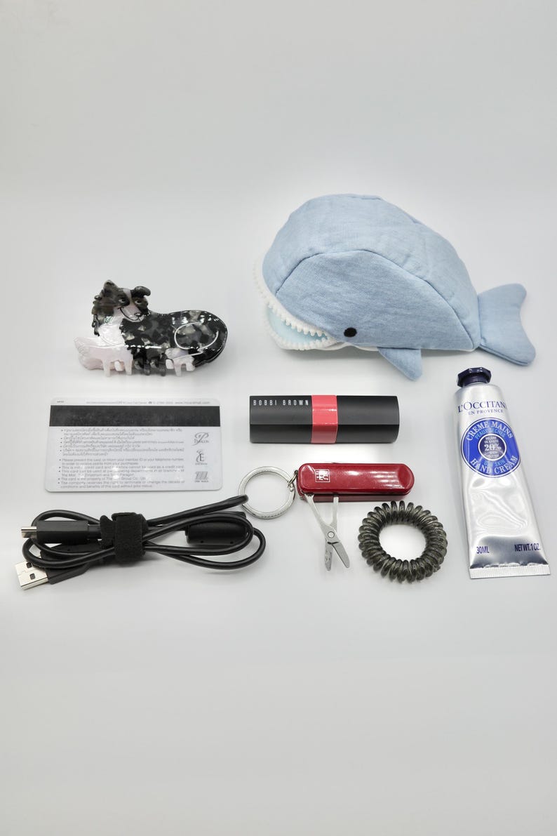May include: A collection of various items including a black and white dog figurine, a blue whale plush toy, a black credit card, a black USB cable, a red lipstick, a red multi-tool keychain, a black hair tie, and a blue tube of hand cream labelled "L'OCCITANE CREME MAINS HAND CREAM".