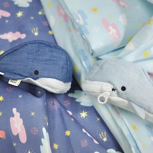 May include: Two blue whale-shaped zippered pouches with white accents. The pouches are made of denim fabric and have a cute, cartoon-like design. The pouches are lying on a blue fabric with a white star and cloud pattern.