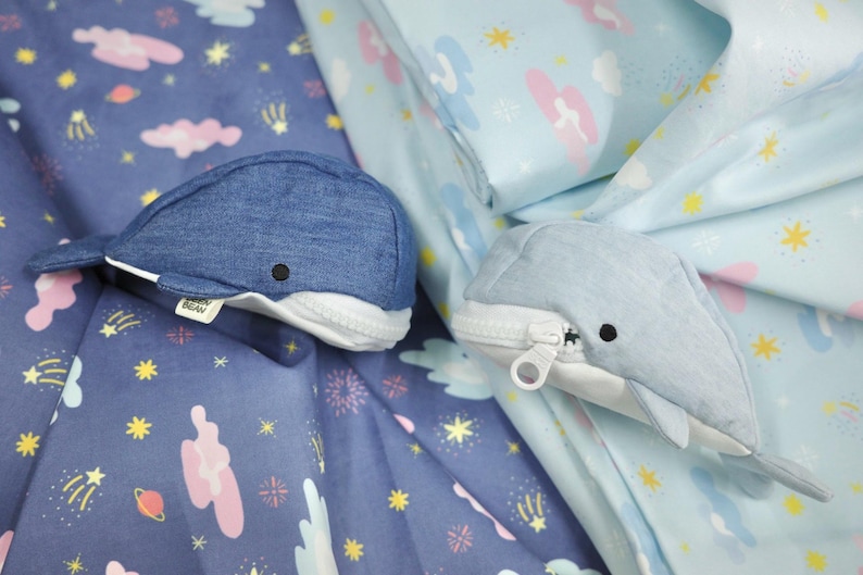 May include: Two blue whale-shaped zippered pouches with white accents. The pouches are made of denim fabric and have a cute, cartoon-like design. The pouches are lying on a blue fabric with a white star and cloud pattern.