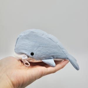 May include: A light blue whale-shaped zippered pouch with a white zipper pull. The pouch is made of soft fabric and has a cute whale face.