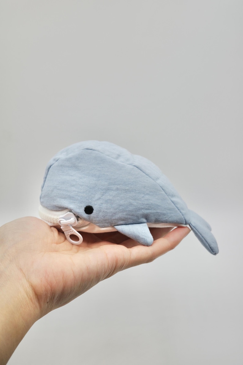 May include: A light blue whale-shaped zippered pouch with a white zipper pull. The pouch is made of soft fabric and has a cute whale face.