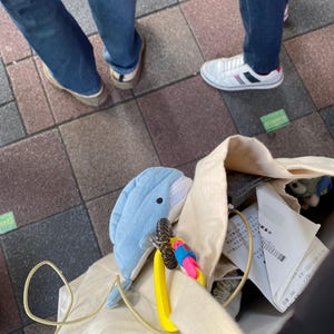 May include: A white canvas tote bag with a blue dolphin plush toy attached to a yellow carabiner. The bag is filled with various items, including a receipt and a grey fabric item.