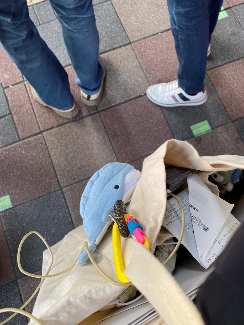 May include: A white canvas tote bag with a blue dolphin plush toy attached to a yellow carabiner. The bag is filled with various items, including a receipt and a grey fabric item.