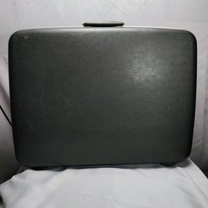 May include: A rectangular black hard-shell suitcase with a textured surface and rounded corners. The suitcase features a centered black top handle and a smooth, solid exterior design, providing a clean and functional appearance for travel.