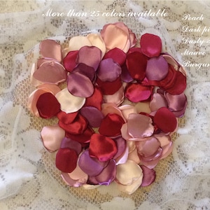 May include: A collection of satin rose petals in various shades of pink, red, and purple. The petals are arranged in a heart shape on a white lace background. The text "More than 25 colors available" is visible at the top of the image. The text "Peach, Dark pink, Dusty Rose, Mauve, Burgundy" is visible on the right side of the image.