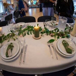 May include: A white tablecloth with green foliage and candles. There are two place settings with white plates, green napkins, and silverware. The table is set for a formal dinner.