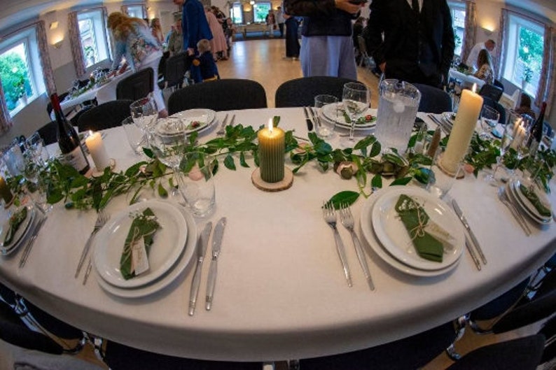 May include: A white tablecloth with green foliage and candles. There are two place settings with white plates, green napkins, and silverware. The table is set for a formal dinner.