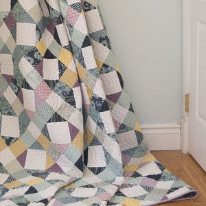 May include: A quilt with a geometric pattern in shades of blue, green, yellow, and purple. The quilt is folded and draped over a corner of a room.