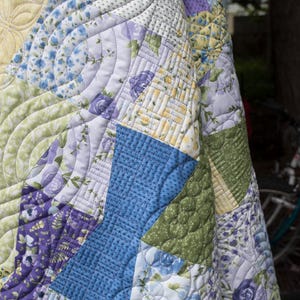 Op de afbeelding: Een patchwork quilt met verschillende bloempatronen in blauw, paars, groen en geel. De quilt heeft ingewikkelde gewatteerde ontwerpen, waaronder gebogen lijnen en bloemmotieven, waardoor een textiel ontstaat met textuur en visuele aantrekkingskracht.
