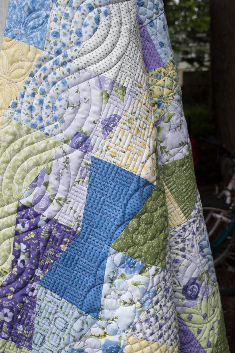 Op de afbeelding: Een patchwork quilt met verschillende bloempatronen in blauw, paars, groen en geel. De quilt heeft ingewikkelde gewatteerde ontwerpen, waaronder gebogen lijnen en bloemmotieven, waardoor een textiel ontstaat met textuur en visuele aantrekkingskracht.