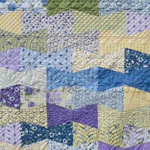 Op de afbeelding: Close-up van een patchworkquilt met een verscheidenheid aan patronen en kleuren. De quilt heeft vierkanten en geometrische vormen in tinten blauw, geel, groen en paars. De quiltsteken voegen textuur en detail toe aan het handgemaakte textiel.