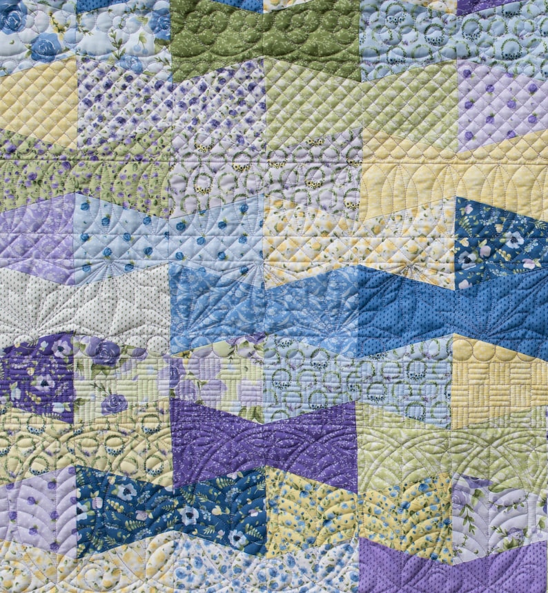 Op de afbeelding: Close-up van een patchworkquilt met een verscheidenheid aan patronen en kleuren. De quilt heeft vierkanten en geometrische vormen in tinten blauw, geel, groen en paars. De quiltsteken voegen textuur en detail toe aan het handgemaakte textiel.