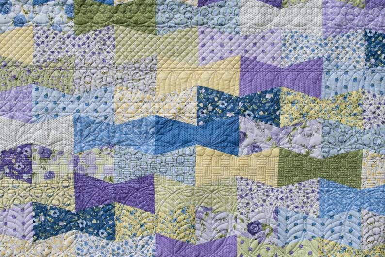Op de afbeelding: Close-up van een patchwork quilt met een verscheidenheid aan patronen en kleuren. De quilt heeft geometrische vormen in tinten blauw, groen, geel en paars. De quiltsteken voegen textuur en detail toe aan de stof.