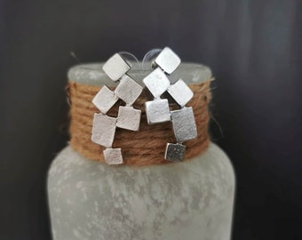 May include: A pair of silver earrings with a geometric design. The earrings are made of multiple small squares that are connected together. The earrings are hanging from a brown rope.