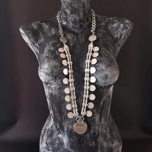 May include: Silver-toned necklace with a coin pendant and multiple disc-shaped charms. The necklace is displayed on a black mannequin. The design features a central pendant with a spiral design and coin-like charms.