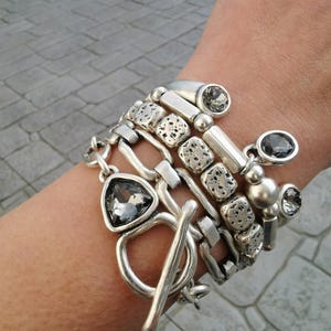 Op de afbeelding: Een stapel zilveren armbanden met verschillende ontwerpen. De armbanden bevatten schakels, getextureerde kralen en geometrische bedels. Verschillende armbanden hebben grijze edelsteenaccenten. De stapel is vastgezet met een tuimelsluiting.