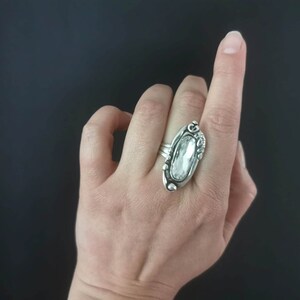 A silver plated ring with a clear oval-shaped stone set in a detailed design. The ring is worn on a finger.
