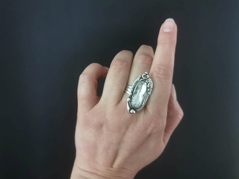 A silver plated ring with a clear oval-shaped stone set in a detailed design. The ring is worn on a finger.