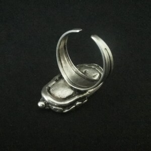 May include: A silver ring with a textured, oval-shaped design. The ring is open on the back and has a smooth, polished finish.
