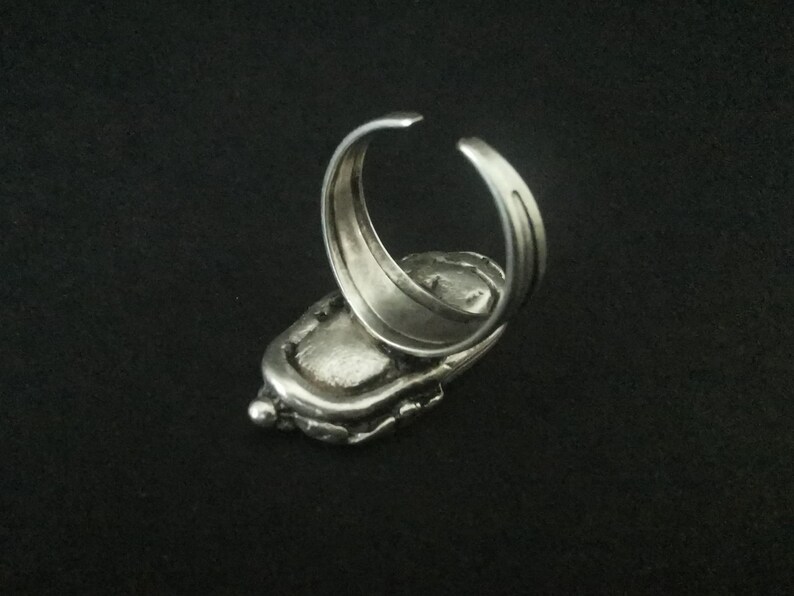 May include: A silver ring with a textured, oval-shaped design. The ring is open on the back and has a smooth, polished finish.