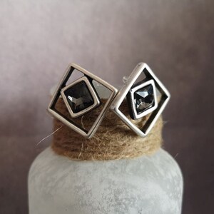 May include: A pair of silver earrings with a geometric design. Each earring features a square frame with a black crystal in the center.