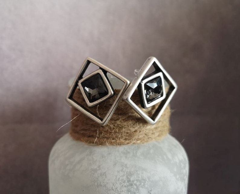 May include: A pair of silver earrings with a geometric design. Each earring features a square frame with a black crystal in the center.