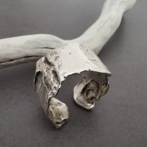 Op de afbeelding: Een zilverkleurige manchetarmband met een textuurrijk, organisch ontwerp. De open manchet heeft een onregelmatige, gebeeldhouwde vorm, met een ruw, ongelijkmatig oppervlak. De armband wordt getoond tegen een grijze achtergrond met een witte tak.