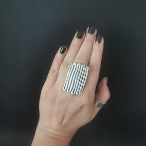 May include: A silver ring with a unique design of multiple stacked, wavy bands. The ring is worn on a hand with black nail polish.