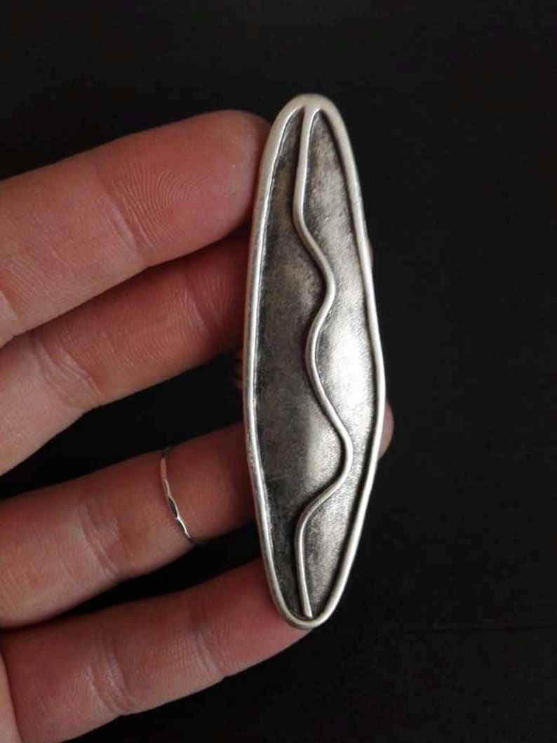 May include: A silver-toned pendant with a wavy line design inside a rounded oval shape.