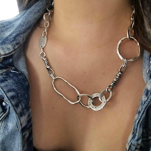 Op de afbeelding: Een zilverkleurige ketting met verschillende schakels in diverse vormen, waaronder cirkels, ovalen en getextureerde elementen. De ketting wordt gedragen tegen een spijkerjasje.