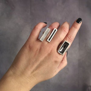 Può includere: Tre anelli d'argento con pietre rettangolari. L'anello sull'indice ha una pietra trasparente, l'anello sul medio ha una pietra nera e l'anello sull'anulare ha una pietra trasparente.