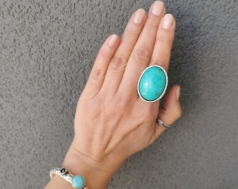 May include: A large oval turquoise ring with a silver band is shown. A silver bracelet with turquoise and black details is also visible. The jewellery is set against a grey background.