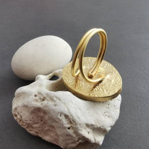 Peut inclure: Une bague dor&eacute;e avec une face ovale textur&eacute;e et une boucle &agrave; double bande. La bague est pos&eacute;e sur une pierre blanche poreuse, avec une pierre ovale blanche lisse en arri&egrave;re-plan. Le motif de la bague pr&eacute;sente un motif floral en relief.