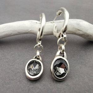 May include: Silver-coloured hoop earrings with a dangling, oval-shaped, grey gemstone. The earrings are displayed against a grey background, showcasing their design and details. The gemstones have a faceted cut, reflecting light.