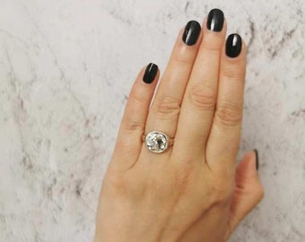 Puede incluir: Un anillo de plata con una gran piedra preciosa transparente de forma cojín. El anillo está en un dedo con las uñas pintadas de negro.